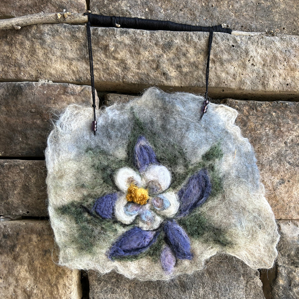 Wool felted blue columbine flower on cream background, with raw wool edges.  Attached to a stick with black thread. Against an outside stone wall.
