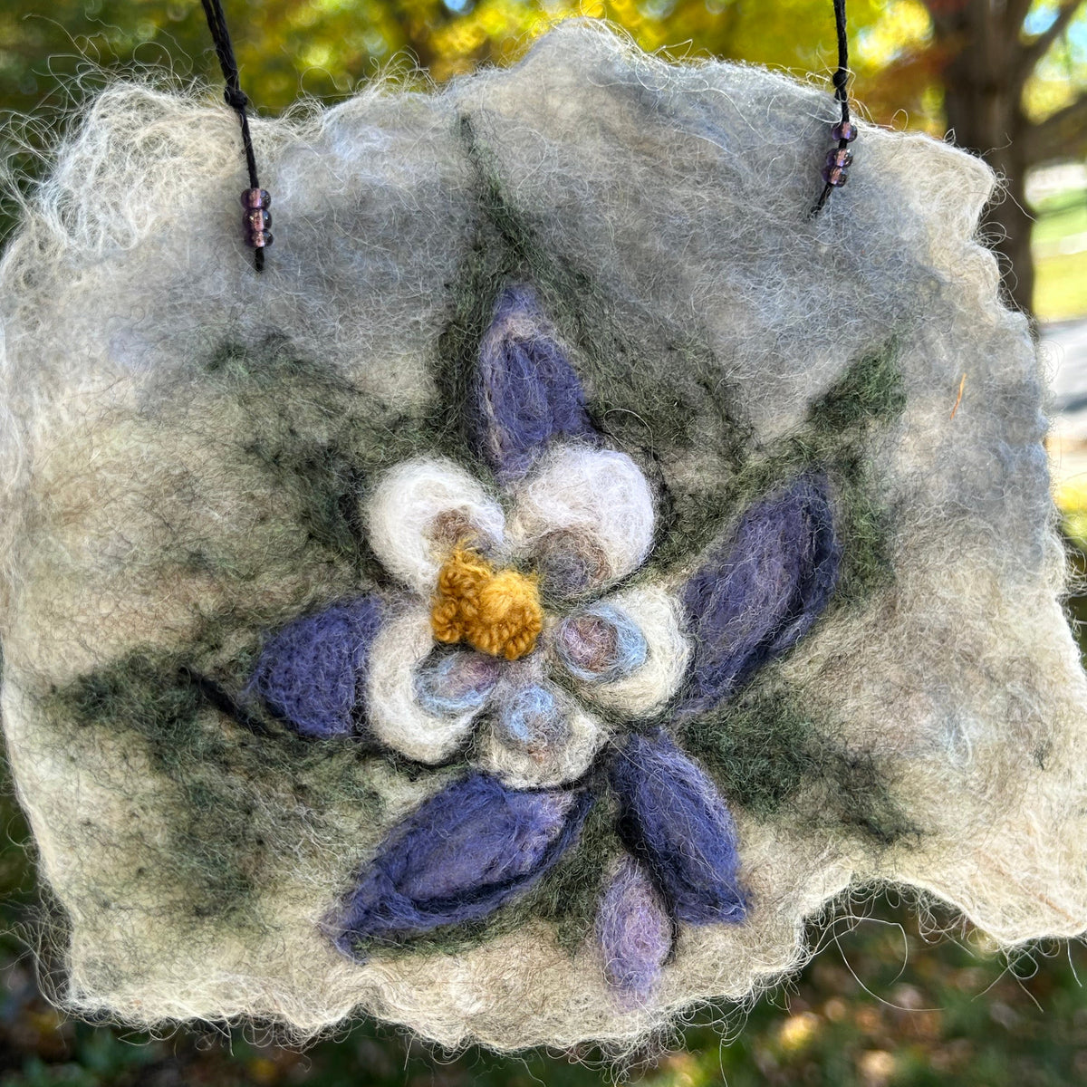 Blue columbine flower wool felted on a cream colored background, outside with a green blurred background
