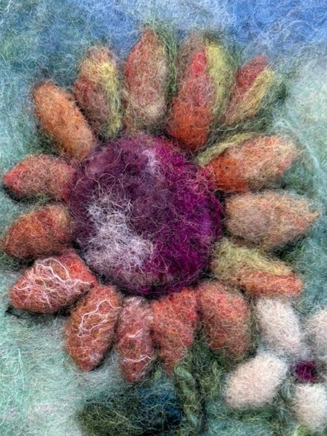 A close-up of a hand-felted wool art piece depicting a large sunflower with orange, pink, and purple petals, green leaves, and a small white flower on a textured blue and green felt background.