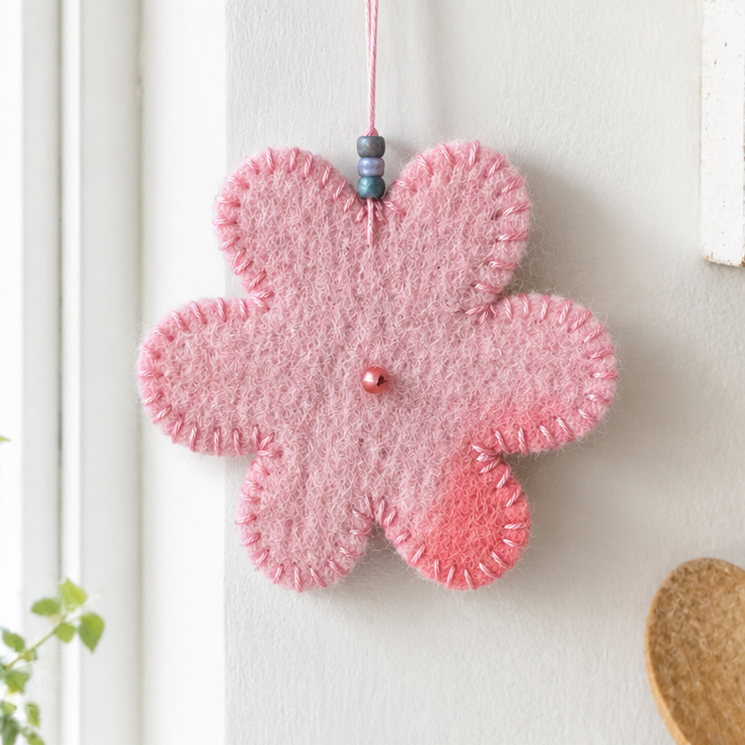 A pink felt flower-shaped ornament hanging from a gold hook on a white wall, with a small bead and string at the top. The ornament has a stitched texture and a small pink bead in the center.