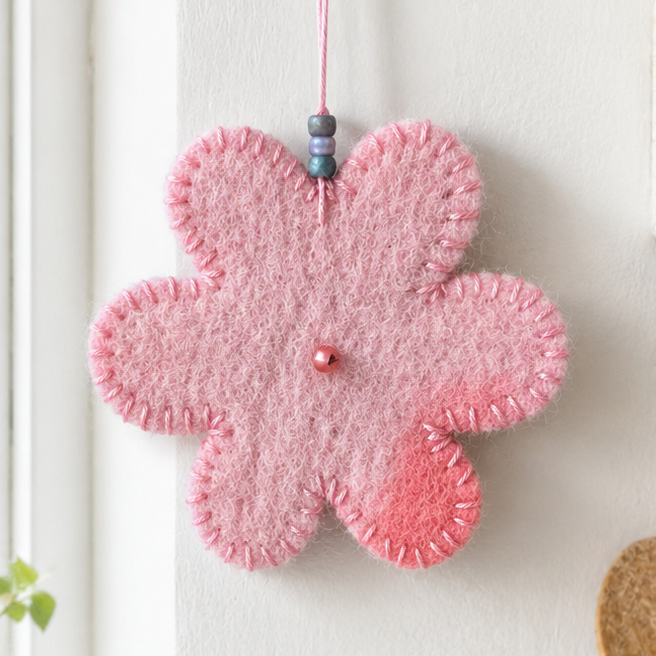 A pink felt flower-shaped ornament hanging from a gold hook on a white wall, with a small bead and string at the top. The ornament has a stitched texture and a small pink bead in the center.