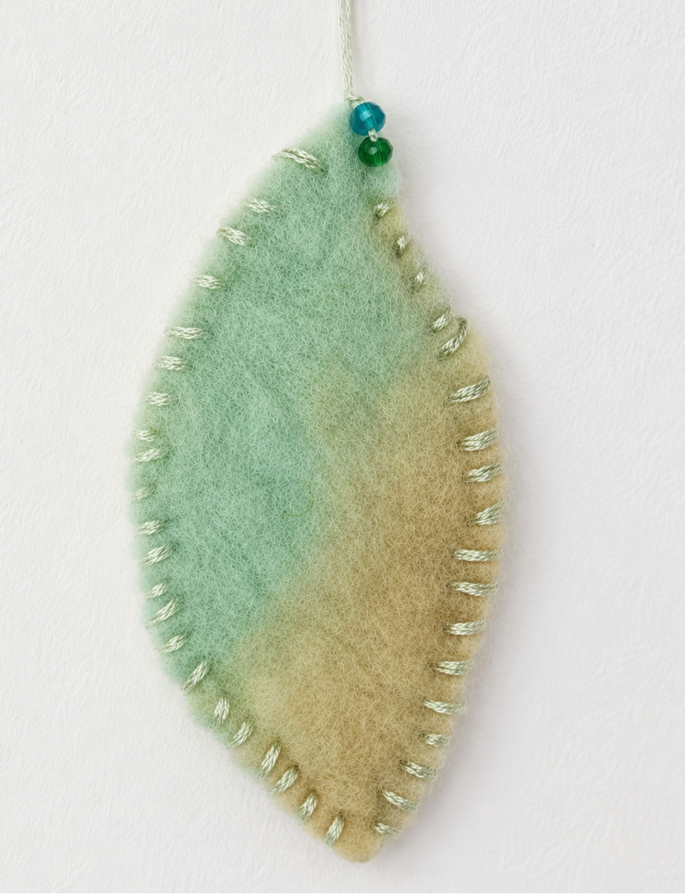 A handcrafted felted wool leaf shape with a green and beige gradient, finished with silver-tone metal beads along the edge and a pin stem with two small blue beads, shown on a plain white background.
