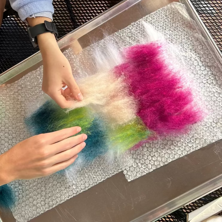Colorful wool layered on a tray. Two hands are applying more to start a wet felted design.