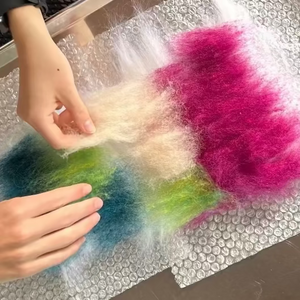 Colorful wool layered on a tray. Two hands are applying more to start a wet felted design.